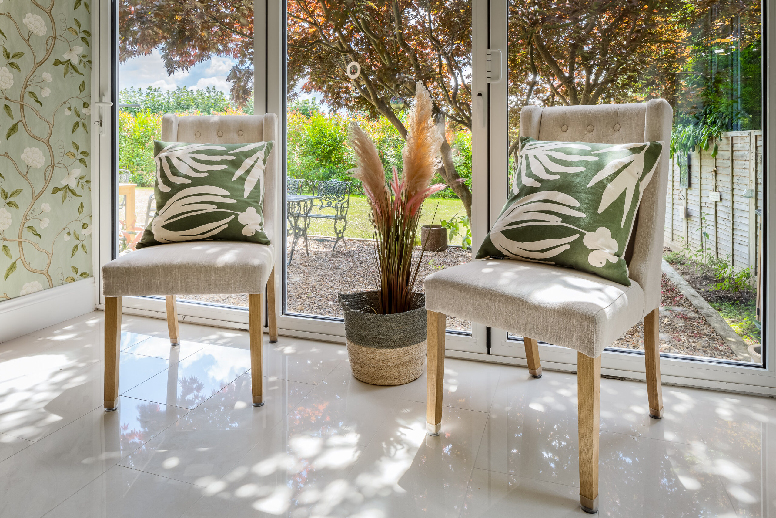 Linen dining chairs with bold botanical green cushions beside garden doors, pampas grass in wicker pot and sage floral wallpaper — biophilic interior design Penn Buckinghamshire