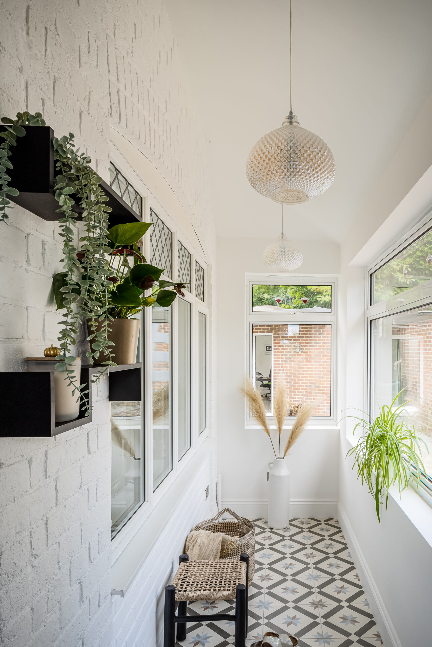 Light-filled garden hallway with encaustic star-pattern floor tiles, white painted brick wall, trailing houseplants on black shelves, pampas grass and diamond pendant lights — biophilic interior design Princes Risborough Buckinghamshire
